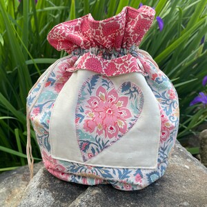 May include: A drawstring bag made of fabric with a floral pattern. The bag has a large heart-shaped patch sewn on the front, also with a floral pattern. The heart is in shades of pink, blue, and green.