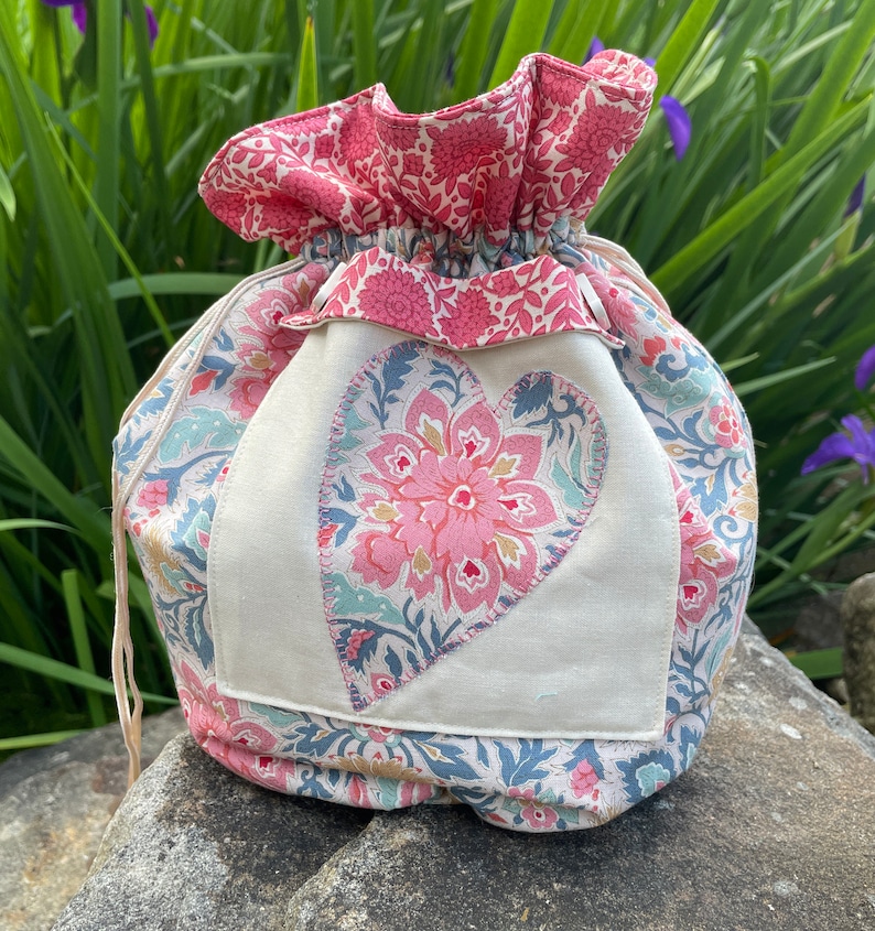 May include: A drawstring bag made of fabric with a floral pattern. The bag has a large heart-shaped patch sewn on the front, also with a floral pattern. The heart is in shades of pink, blue, and green.