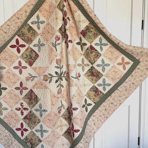 May include: A handmade quilt with a floral and geometric pattern. The quilt features squares in shades of pink, cream, and green, with floral and cross-shaped designs. The border has a floral pattern. The quilt is a traditional craft item.