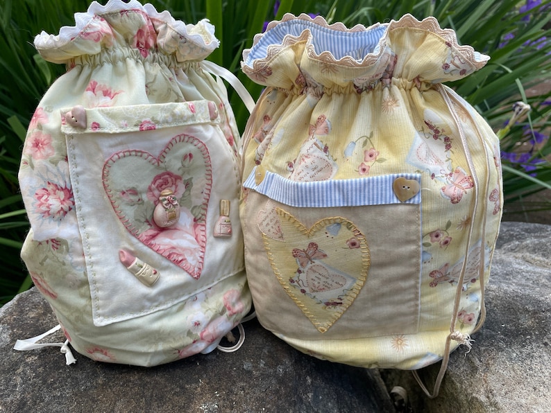 May include: Two drawstring bags made of fabric with floral patterns. Both bags have a heart-shaped patch on the front. One bag is yellow with a light blue stripe and a brown button. The other bag is white with a pink heart and a pink button.