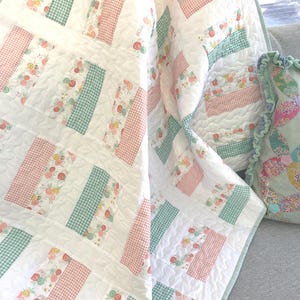 May include: A white quilt with a patchwork design featuring sections of floral patterns, pink gingham, and green gingham. The quilt is folded over a gray surface, with a matching pillow visible.