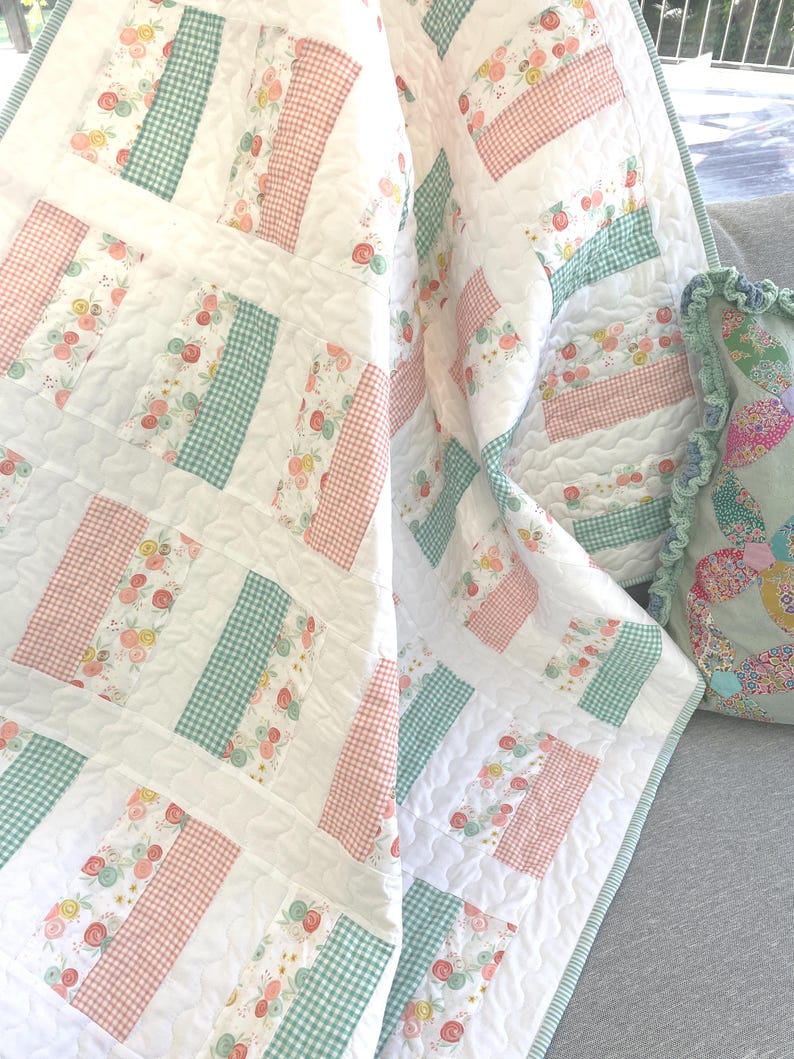 May include: A white quilt with a patchwork design featuring sections of floral patterns, pink gingham, and green gingham. The quilt is folded over a gray surface, with a matching pillow visible.