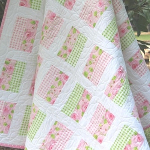 May include: A quilted blanket featuring a repeating pattern of rectangular blocks. Each block consists of a pink floral print section paired with a light green and white gingham section, separated by white fabric strips with wavy stitching.