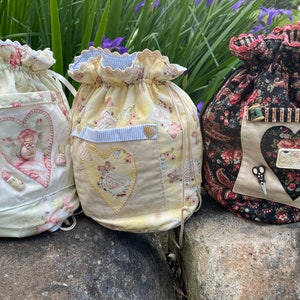 May include: Three drawstring bags with floral patterns and heart-shaped pockets. The bags are made of fabric and have a vintage aesthetic. The bag on the right has a tag that says "Quilt Shop".