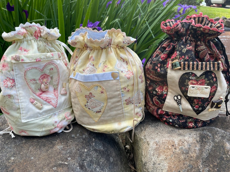 May include: Three drawstring bags with floral patterns and heart-shaped pockets. The bags are made of fabric and have a vintage aesthetic. The bag on the right has a tag that says "Quilt Shop".