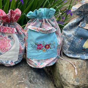 May include: Three fabric drawstring bags with different patterns. The first bag is pink and white with a heart-shaped design. The second bag is blue and white with a floral embroidery. The third bag is blue and white with a woman holding an umbrella.