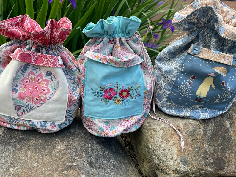 May include: Three fabric drawstring bags with different patterns. The first bag is pink and white with a heart-shaped design. The second bag is blue and white with a floral embroidery. The third bag is blue and white with a woman holding an umbrella.