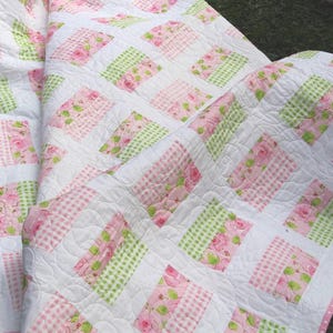 May include: A quilted blanket featuring a repeating pattern of white squares interspersed with rectangular blocks of pink floral fabric, light green gingham, and pink gingham. The quilt is finished with a thin pink border with white dots.