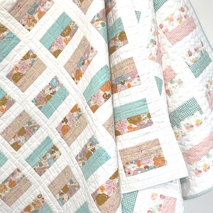 May include: A handmade quilt with a white background and a patchwork design. The quilt features rectangular blocks in shades of blue, tan, and floral patterns. The quilt is made of soft fabric and has a quilted texture.