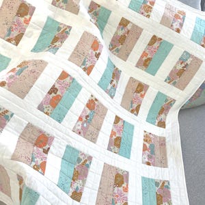 May include: A handmade quilt with a repeating pattern of rectangular blocks. The blocks alternate between floral prints in pink, peach, and brown, and solid light blue sections, all set against a white background. The quilt is made of soft fabric.