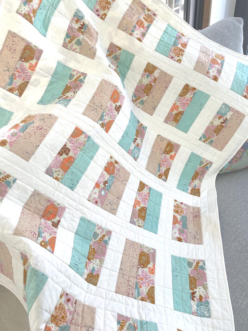 May include: A handmade quilt with a repeating pattern of rectangular blocks. The blocks alternate between floral prints in pink, peach, and brown, and solid light blue sections, all set against a white background. The quilt is made of soft fabric.