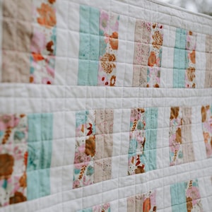 May include: A white quilt with a patchwork design. The quilt features vertical stripes of floral and solid fabric in shades of pink, brown, and teal. The quilt is made of soft fabric and has a quilted pattern.
