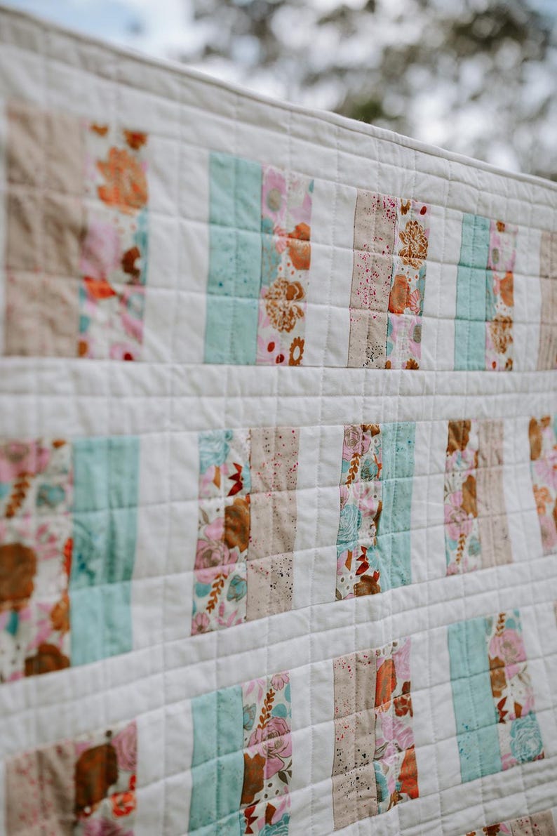 May include: A white quilt with a patchwork design. The quilt features vertical stripes of floral and solid fabric in shades of pink, brown, and teal. The quilt is made of soft fabric and has a quilted pattern.