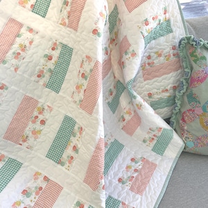 May include: A white quilt with a patchwork design featuring sections of floral patterns, pink gingham, and green gingham. The quilt is quilted with a wavy pattern. A matching pillow with a ruffled edge is also visible.