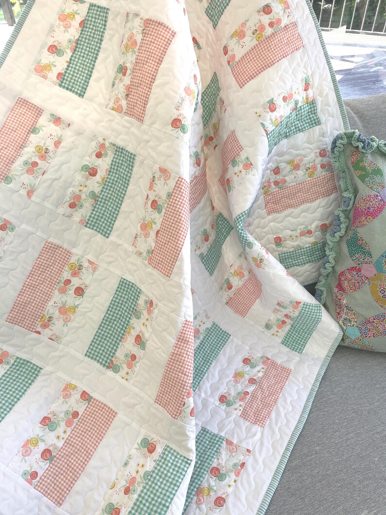 May include: A white quilt with a patchwork design featuring sections of floral patterns, pink gingham, and green gingham. The quilt is quilted with a wavy pattern. A matching pillow with a ruffled edge is also visible.