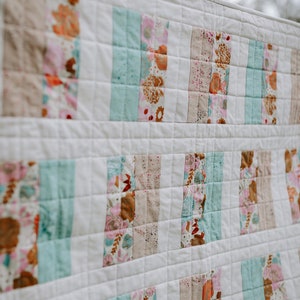 May include: A white quilt with a patchwork design featuring strips of fabric in various shades of pink, brown, and blue. The fabric has a floral pattern.