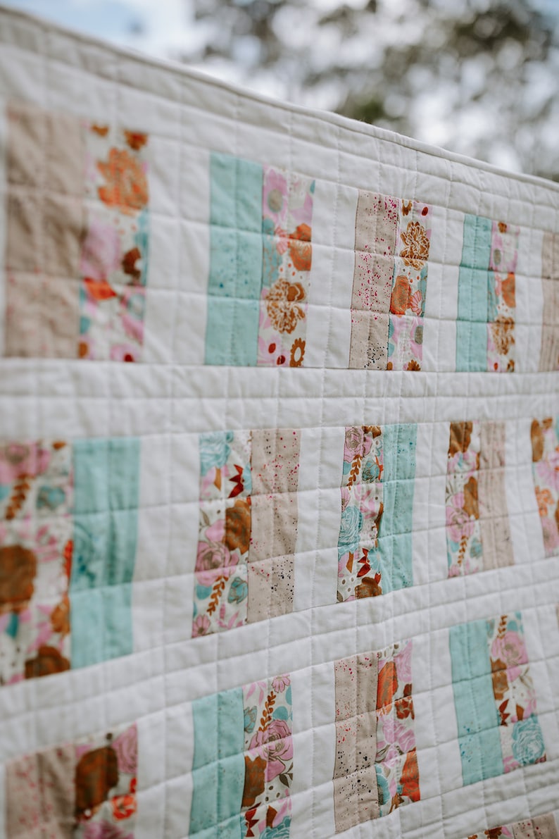 May include: A white quilt with a patchwork design featuring strips of fabric in various shades of pink, brown, and blue. The fabric has a floral pattern.