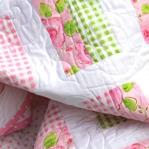 May include: A close-up of a quilted blanket with a patchwork design. The quilt features white, pink, and green squares. Some squares have a pink gingham pattern, others have a floral rose pattern, and some have a green gingham pattern.