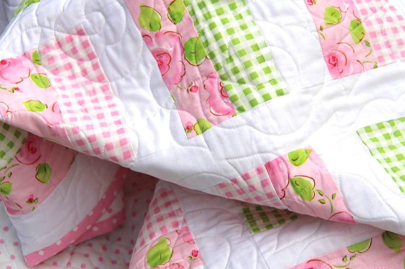 May include: A close-up of a quilted blanket with a patchwork design. The quilt features white, pink, and green squares. Some squares have a pink gingham pattern, others have a floral rose pattern, and some have a green gingham pattern.