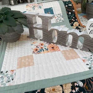 May include: A white quilted table runner with a green border and a floral pattern. The runner has a wooden sign that says "Home" with a heart-shaped sign that says "love the life you live" on top of it.
