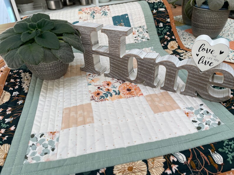 May include: A white quilted table runner with a green border and a floral pattern. The runner has a wooden sign that says "Home" with a heart-shaped sign that says "love the life you live" on top of it.