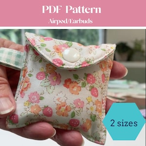 Peut inclure: Une petite pochette en tissu pour Airpods ou écouteurs avec un motif floral et fraises dans des tons rose, orange et jaune sur fond blanc. La pochette possède une fermeture à bouton-pression blanc et une languette latérale à carreaux vert clair.