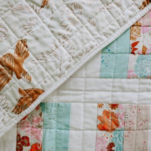 May include: A patchwork quilt with a cream-colored background. One section features brown bird illustrations, while the other has floral and striped patterns in pink, orange, and blue. The quilt is quilted with a diamond pattern.