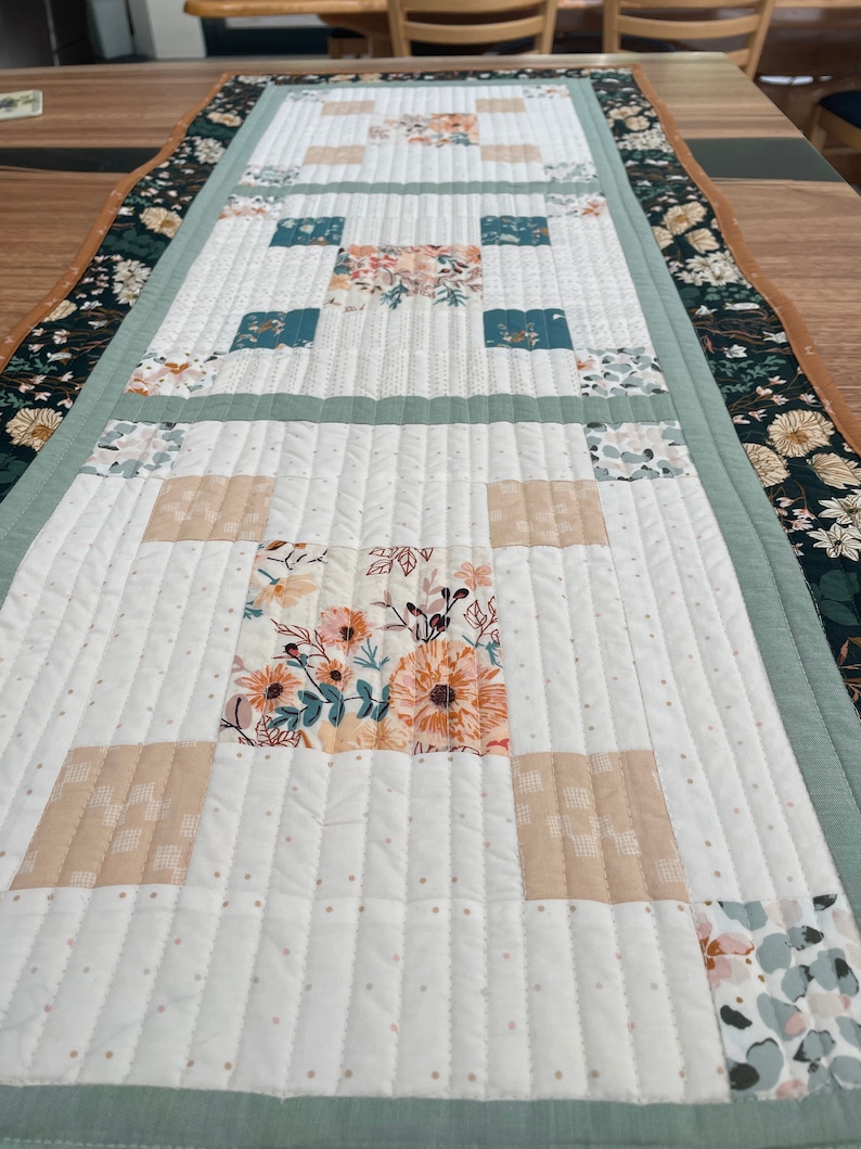 May include: A white table runner with a green border and a floral pattern in the center. The runner is quilted with a small white dot pattern.