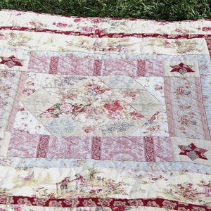 May include: A rectangular quilt featuring a central hexagonal panel with floral patterns in shades of red, pink, and cream. The design includes geometric borders, star-shaped corner accents, and a repeating toile-style print of figures.