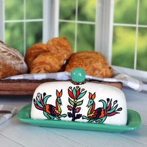 Puede incluir: Una mantequera de cerámica blanca con base verde y pomo turquesa. La tapa presenta coloridos pájaros y diseños florales de arte popular. El plato es rectangular y se asienta sobre una superficie azul claro, con pan al fondo.
