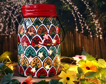 May include: A colorful glass jar with a red lid and a black handle. The jar is decorated with a pattern of green, red, yellow, and orange scales. There are small white stars cut out of the jar, and small white lights are shining through the stars.