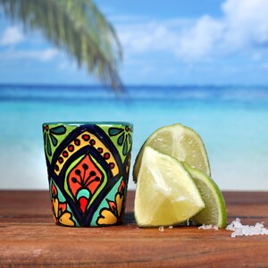 May include: A colorful ceramic shot glass with a geometric pattern in shades of green, blue, yellow, orange, and red.  The shot glass is sitting on a wooden surface with lime wedges and salt.
