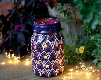 May include: A decorative purple and white mason jar lantern with a star pattern and a metal handle. The jar is illuminated with warm white lights, creating a cozy ambiance. The lantern is placed on a wooden surface.