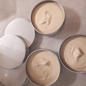 May include: Four round metal containers with lids, filled with a light brown cream-colored substance. The containers are arranged in a semi-circle on a textured surface.