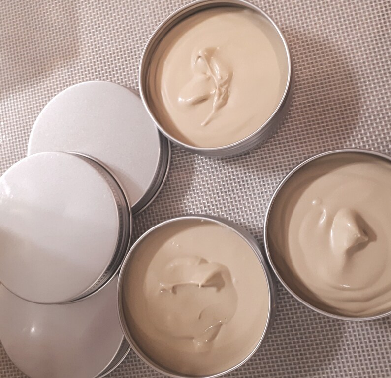 May include: Four round metal containers with lids, filled with a light brown cream-colored substance. The containers are arranged in a semi-circle on a textured surface.