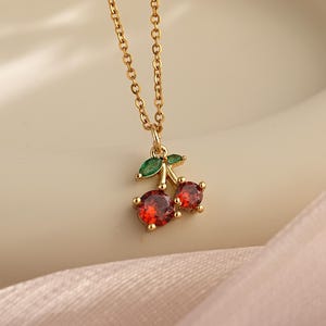 Peut inclure: Un collier en chaîne dorée avec un pendentif cerise rouge et vert. Le pendentif représente deux cerises rouges avec des feuilles vertes.