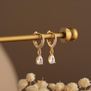 May include: A pair of gold hoop earrings with a teardrop-shaped crystal dangling from each hoop. The hoops are accented with small clear stones.