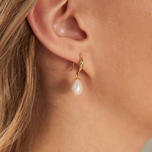 May include: A gold-colored earring with a teardrop-shaped white pearl. The earring is hanging from a person's ear. The earring's hook has a curved design. The pearl is smooth and has a slight luster.