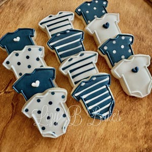 May include: A collection of onesie-shaped sugar cookies decorated with royal icing in navy blue and white. The designs include horizontal stripes, polka dots, and solid colors with small heart accents. The text Lizy B Bakes is visible.
