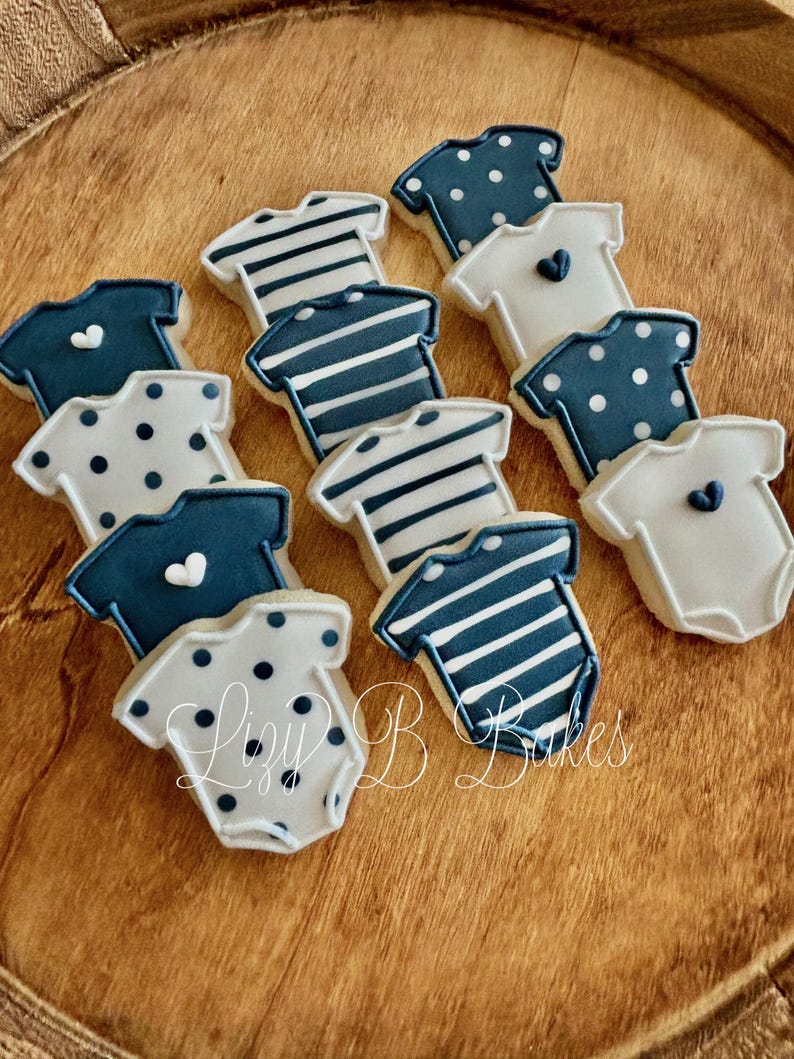 May include: A collection of onesie-shaped sugar cookies decorated with royal icing in navy blue and white. The designs include horizontal stripes, polka dots, and solid colors with small heart accents. The text Lizy B Bakes is visible.