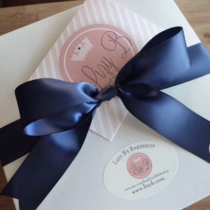 May include: A white gift box with a blue satin ribbon tied in a bow. The box has a pink and white striped card with a logo that says "Lizzy B's Bakeshop" and a website address.