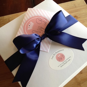 May include: A square white box adorned with a large navy blue satin ribbon tied in a bow. A pink and white striped card with a circular logo reading Lizy B is tucked under the ribbon. An oval sticker on the box reads Lizy B's Bakeshop.