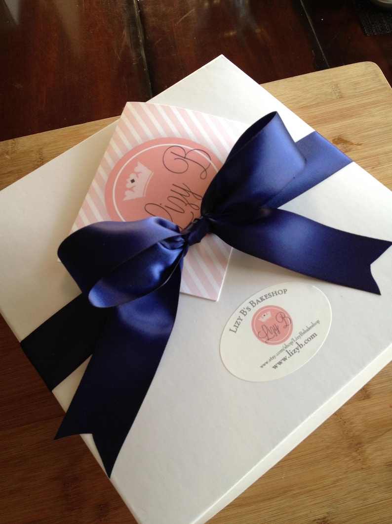 May include: A square white box adorned with a large navy blue satin ribbon tied in a bow. A pink and white striped card with a circular logo reading Lizy B is tucked under the ribbon. An oval sticker on the box reads Lizy B's Bakeshop.