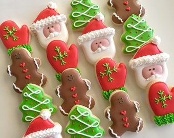 May include: A selection of Christmas biscuits. The biscuits feature Santa faces, red mittens with green snowflake designs, green Christmas trees with white icing, and gingerbread men with heart decorations. The biscuits are decorated with red, white, green, and brown icing.