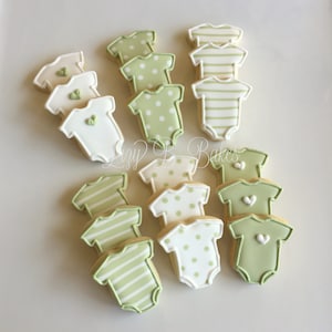 May include: A collection of baby onesie-shaped sugar cookies decorated with icing in shades of sage green, cream, and white. The cookies feature patterns including horizontal stripes, polka dots, and solid colors with small heart accents.