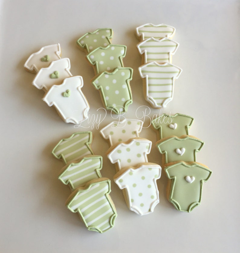 May include: A collection of baby onesie-shaped sugar cookies decorated with icing in shades of sage green, cream, and white. The cookies feature patterns including horizontal stripes, polka dots, and solid colors with small heart accents.