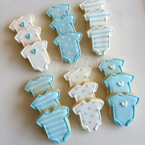 May include: A collection of baby bodysuit-shaped sugar cookies decorated with royal icing. The cookies feature various designs including solid white with blue hearts, light blue with white polka dots, blue stripes, and solid light blue with hearts.