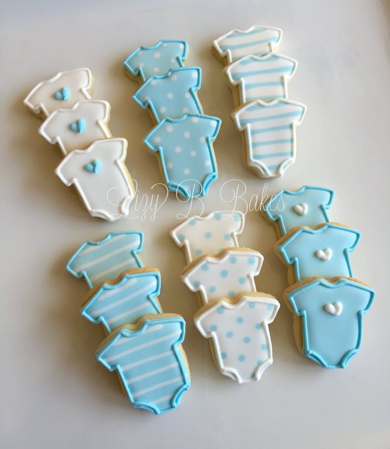 May include: A collection of baby bodysuit-shaped sugar cookies decorated with royal icing. The cookies feature various designs including solid white with blue hearts, light blue with white polka dots, blue stripes, and solid light blue with hearts.
