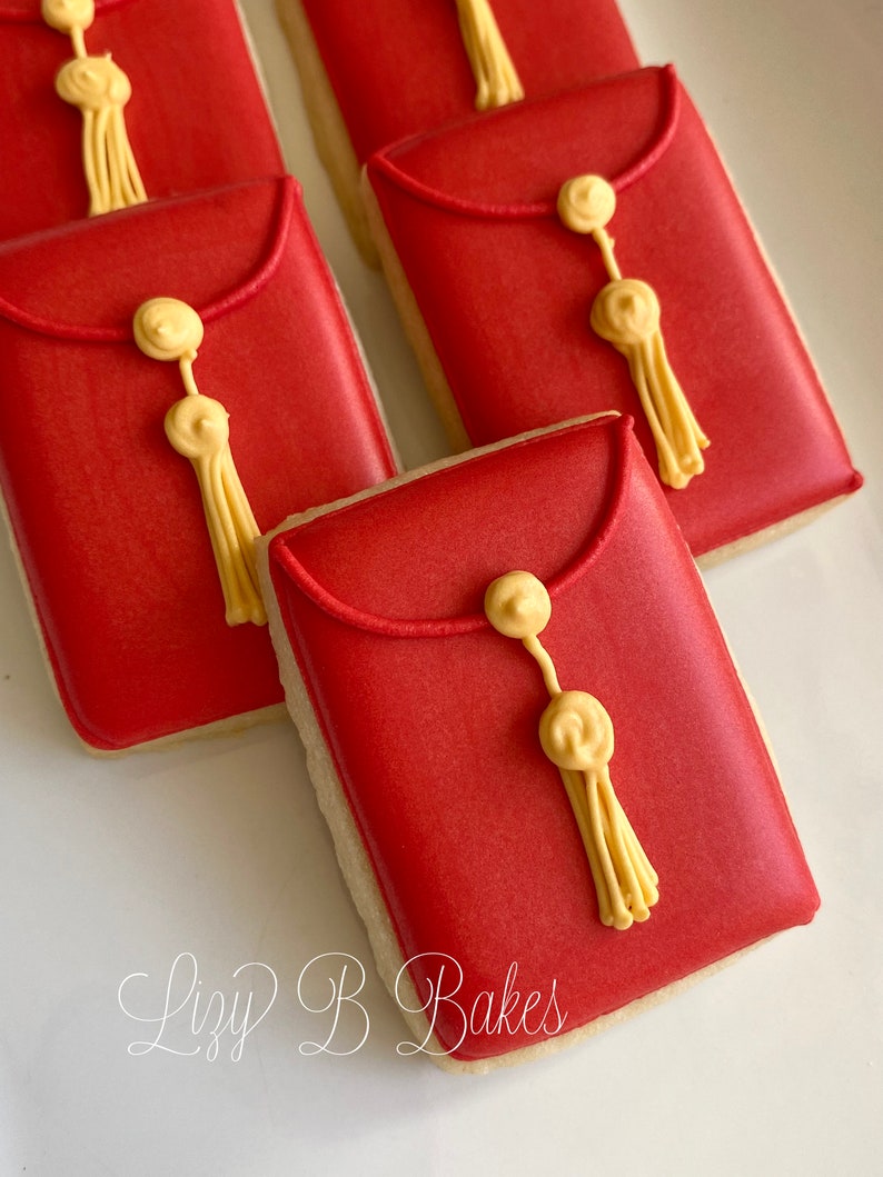May include: Red and gold sugar cookies decorated to look like envelopes with a gold tassel. The cookies are on a white surface with the text "Lizzy B Bakes" written in cursive.