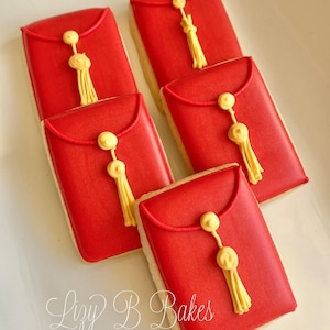 May include: Five red sugar cookies decorated with gold icing to resemble envelopes with tassels. The cookies are arranged in a staggered pattern on a white surface. The text "Lizzy B Bakes" is written in cursive on the surface.